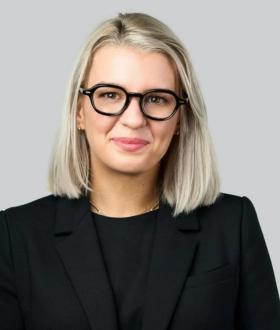 A woman with shoulder-length blonde hair and black glasses smiles subtly. She's wearing a black blazer. The background is a plain light gray.