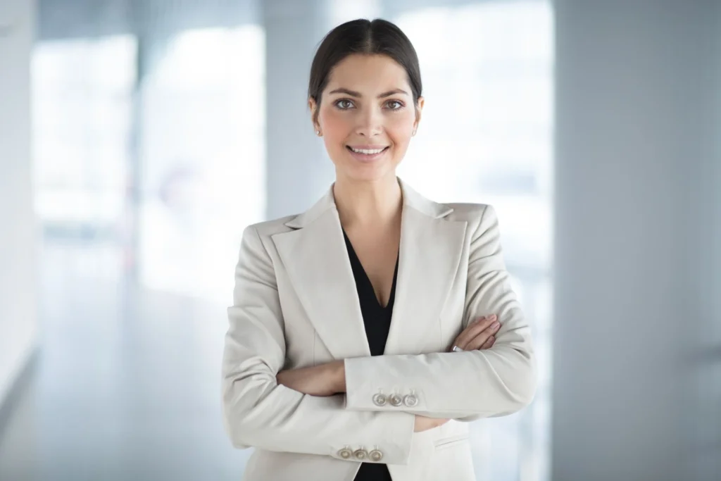 Smiling woman with dark hair in a beige blazer stands confidently with arms crossed. Bright, blurred office background, conveying professionalism.