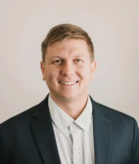 A man in a dark suit and light checkered shirt smiles warmly against a plain background, conveying a friendly and professional demeanor.