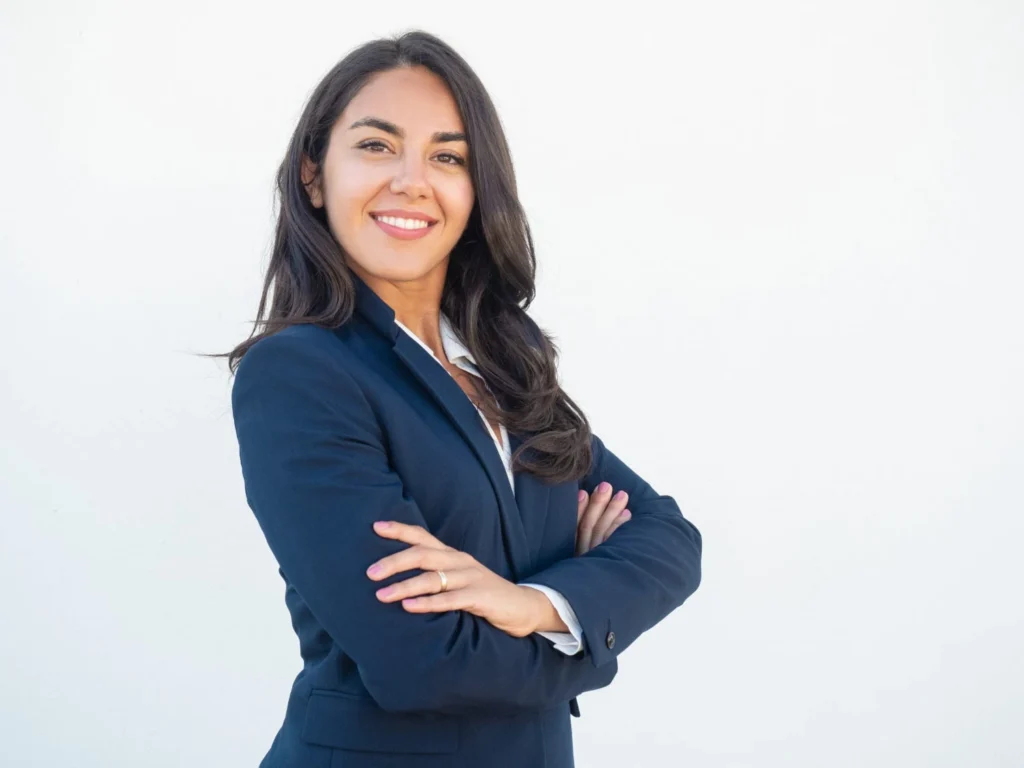 A confident woman in a navy blazer stands with arms crossed against a plain white background, smiling warmly, conveying professionalism and positivity.